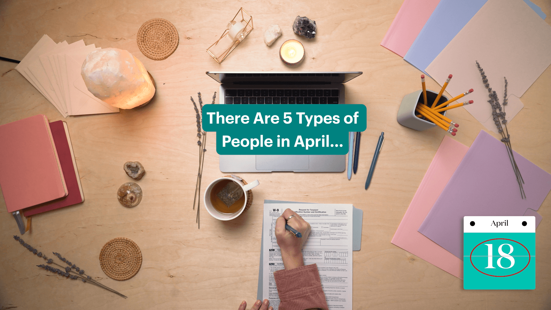 Hand filling out a tax form on a desk with a computer, tea, and assorted desk materials