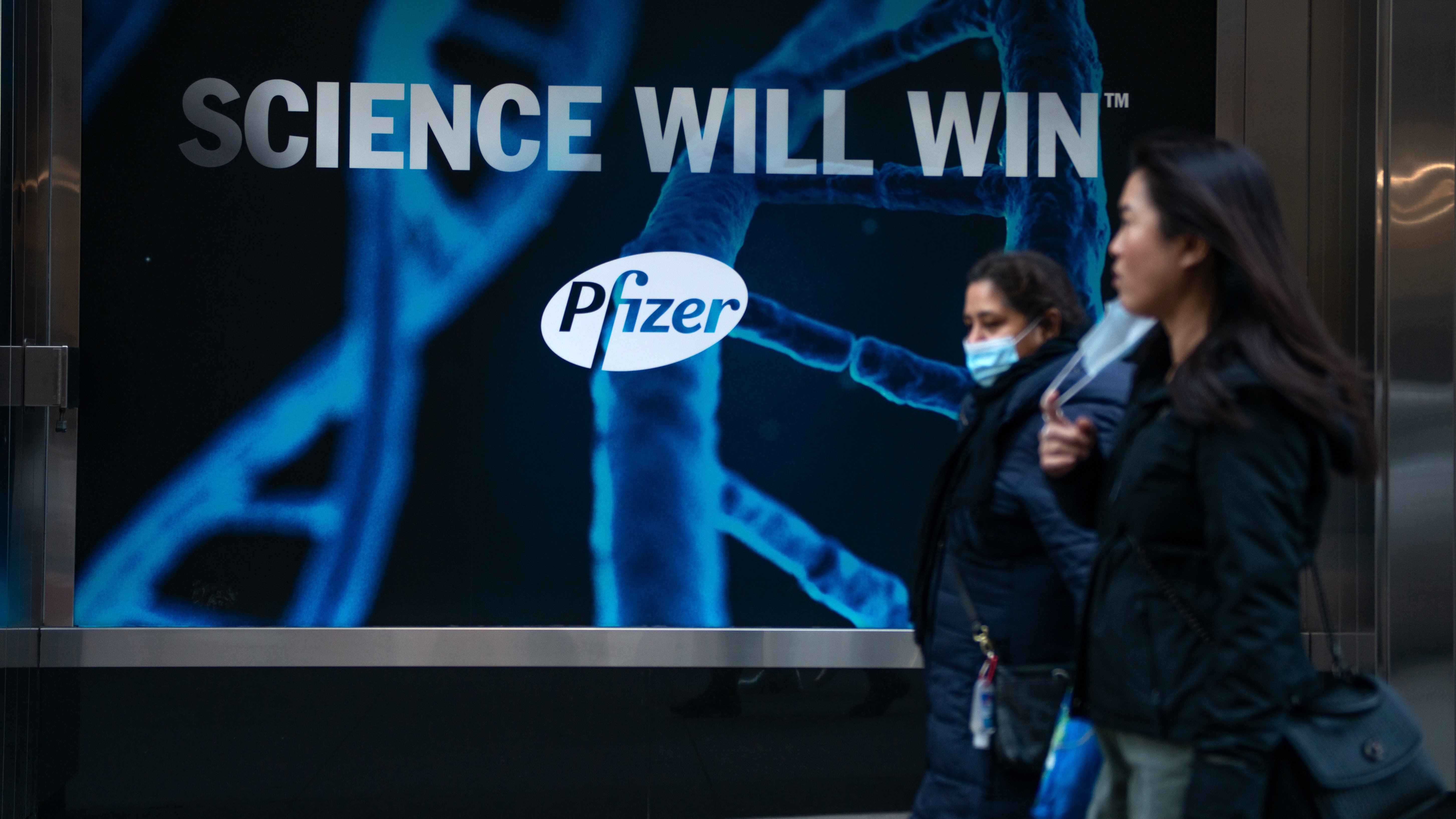 Women with and without a mask walk past a "science will win, Pfizer" sign on December 13, 2020 in New York City.