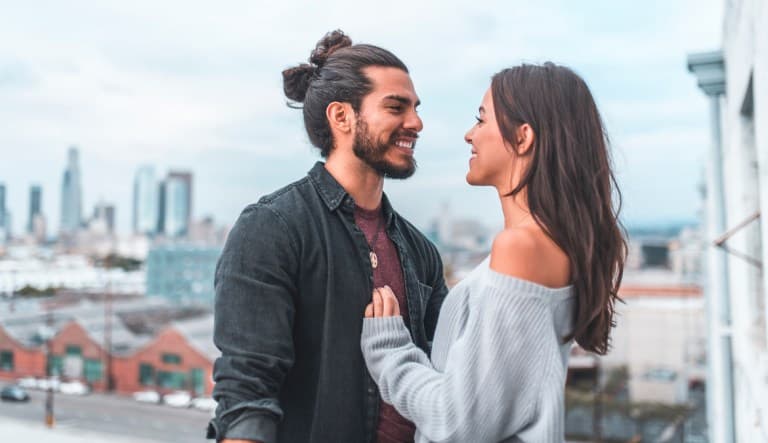 A man and woman smile at each other on the balcony in front of a cityscape. They both have dark hair and are smiling widely. This photo is being used to promote an article about difference between being in love and loving someone.