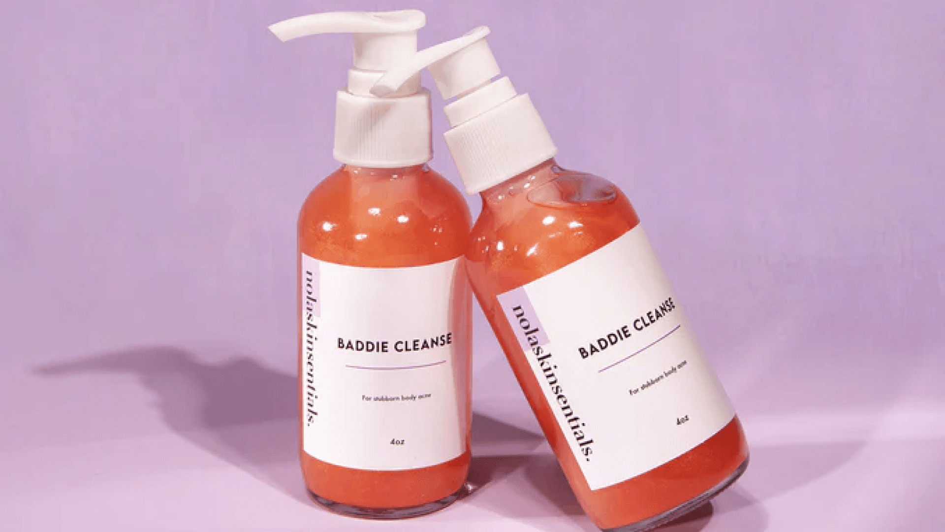 Two bottles of orange body wash leaning against each other resting on a purple background
